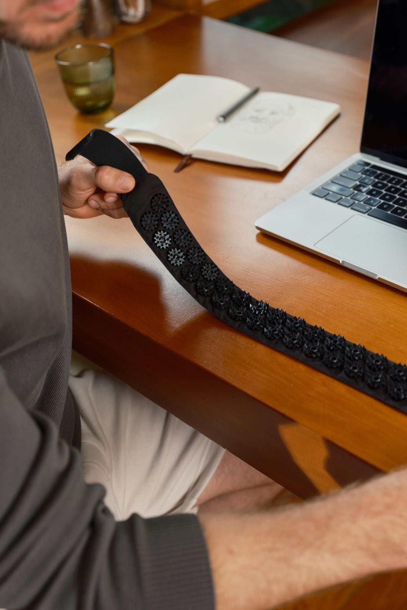 A person holding a Shakti Acupress Headband, shown over a wooden desk with a laptop, notebook, and glass in the background.