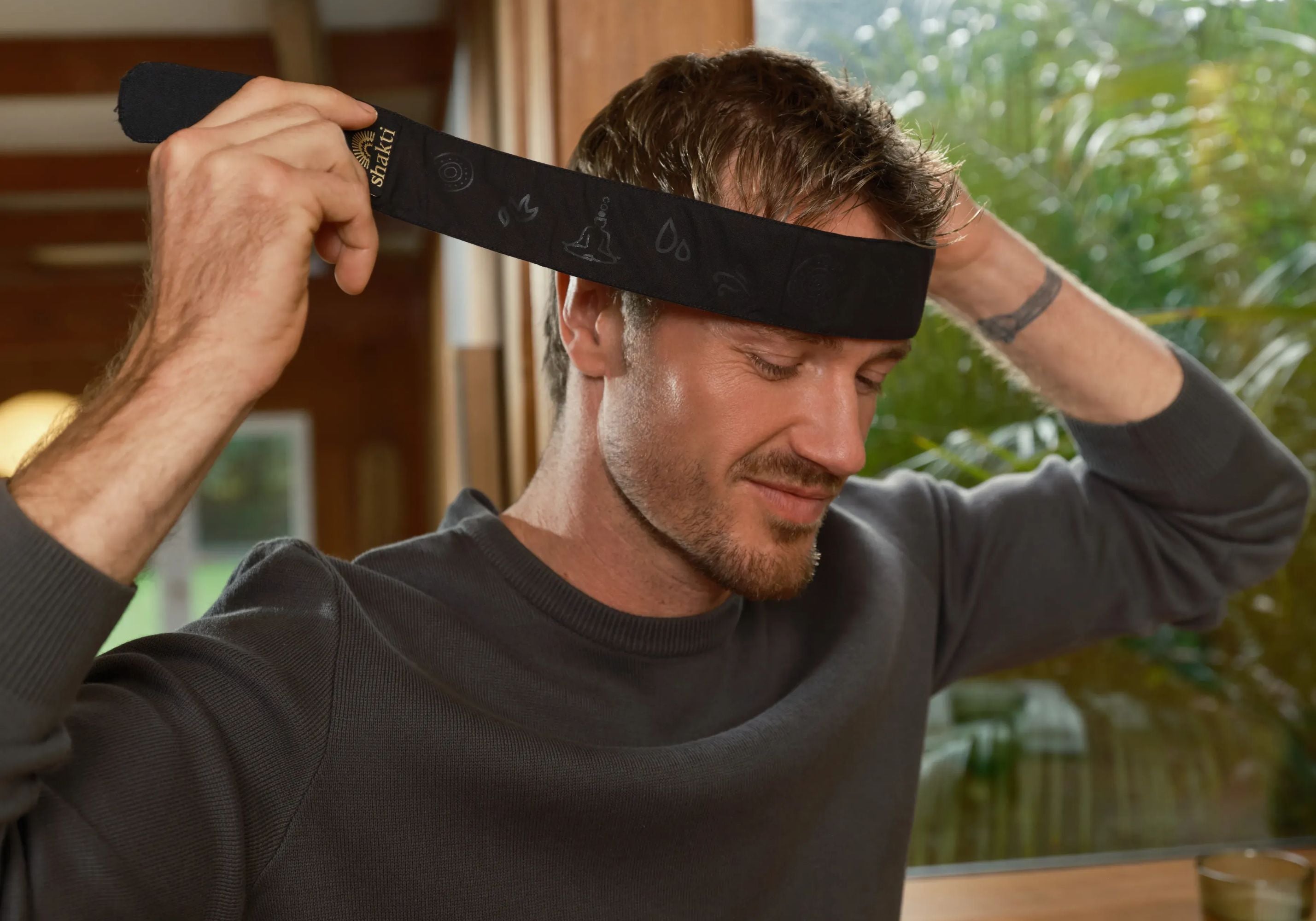 A man wearing a dark gray long-sleeve top adjusting a Acupress Headband with a subtle pattern and a small gold Shakti tag, set against a warm, softly blurred indoor background.