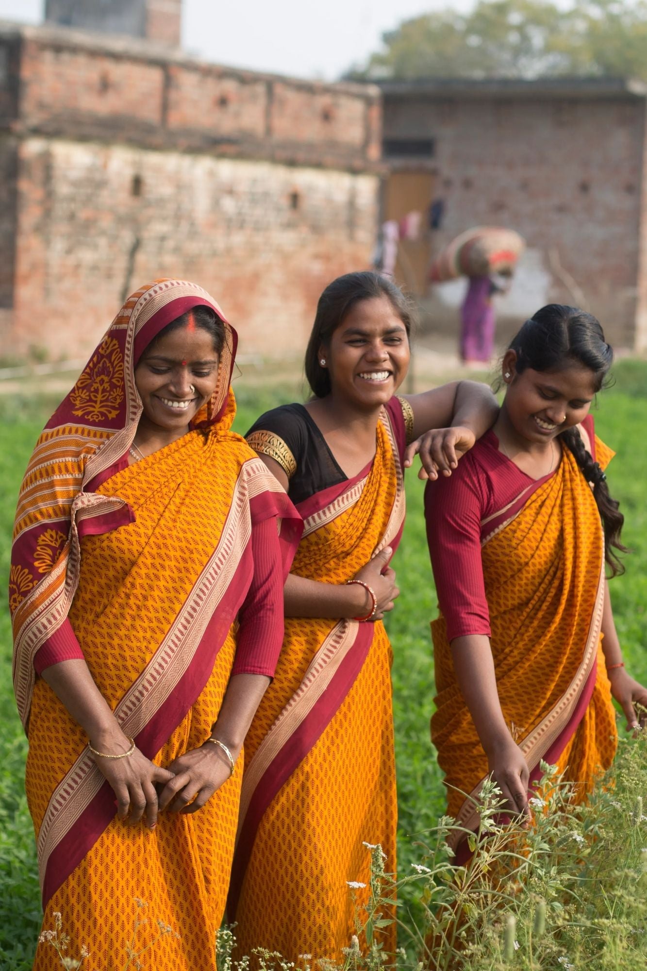 Three women in traditional orange and red sarees smiling and standing in a field.