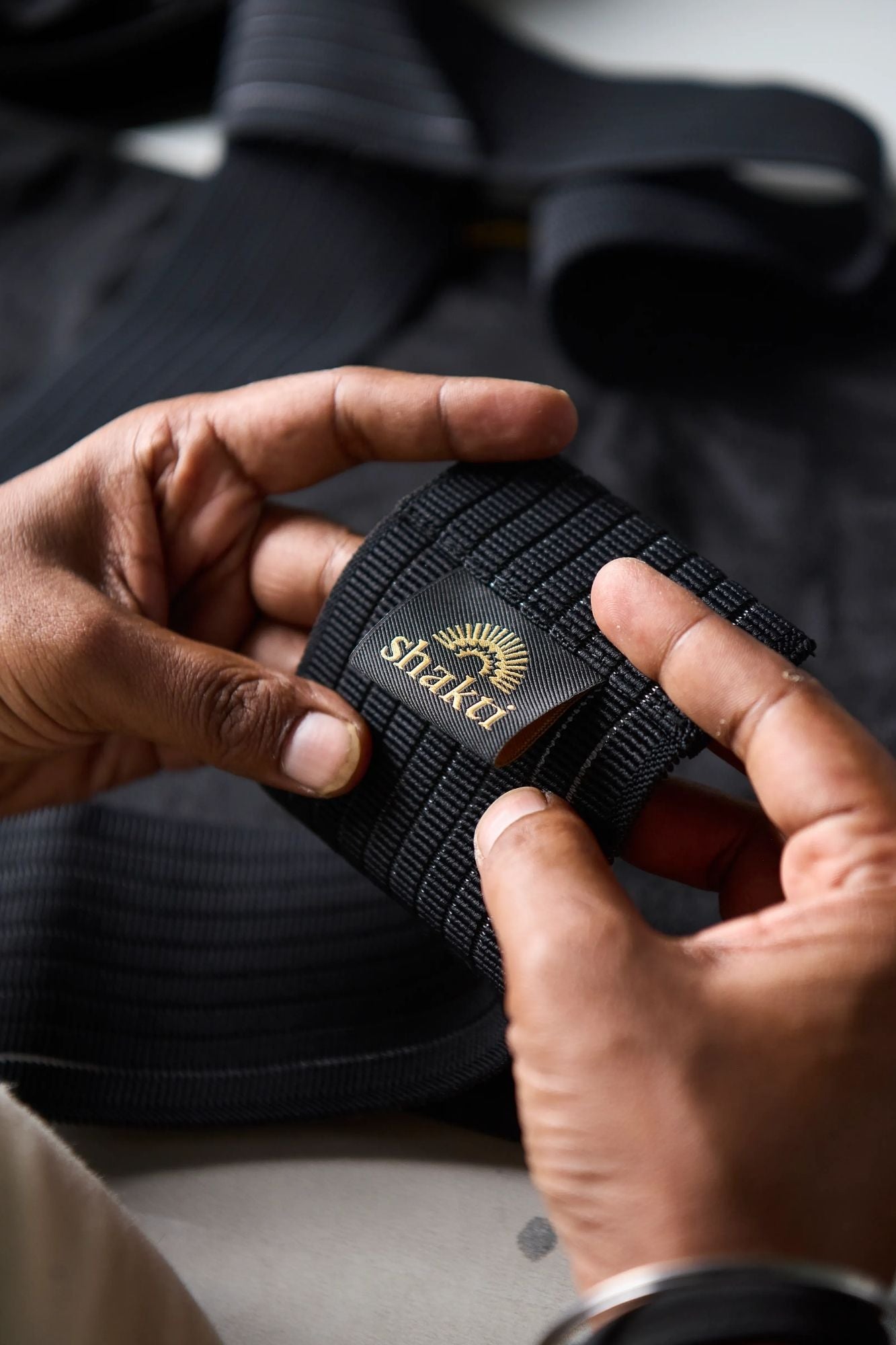 A close-up of a person's hands holding a black ribbed Leg Wrap strap with a gold and black Shakti logo patch.