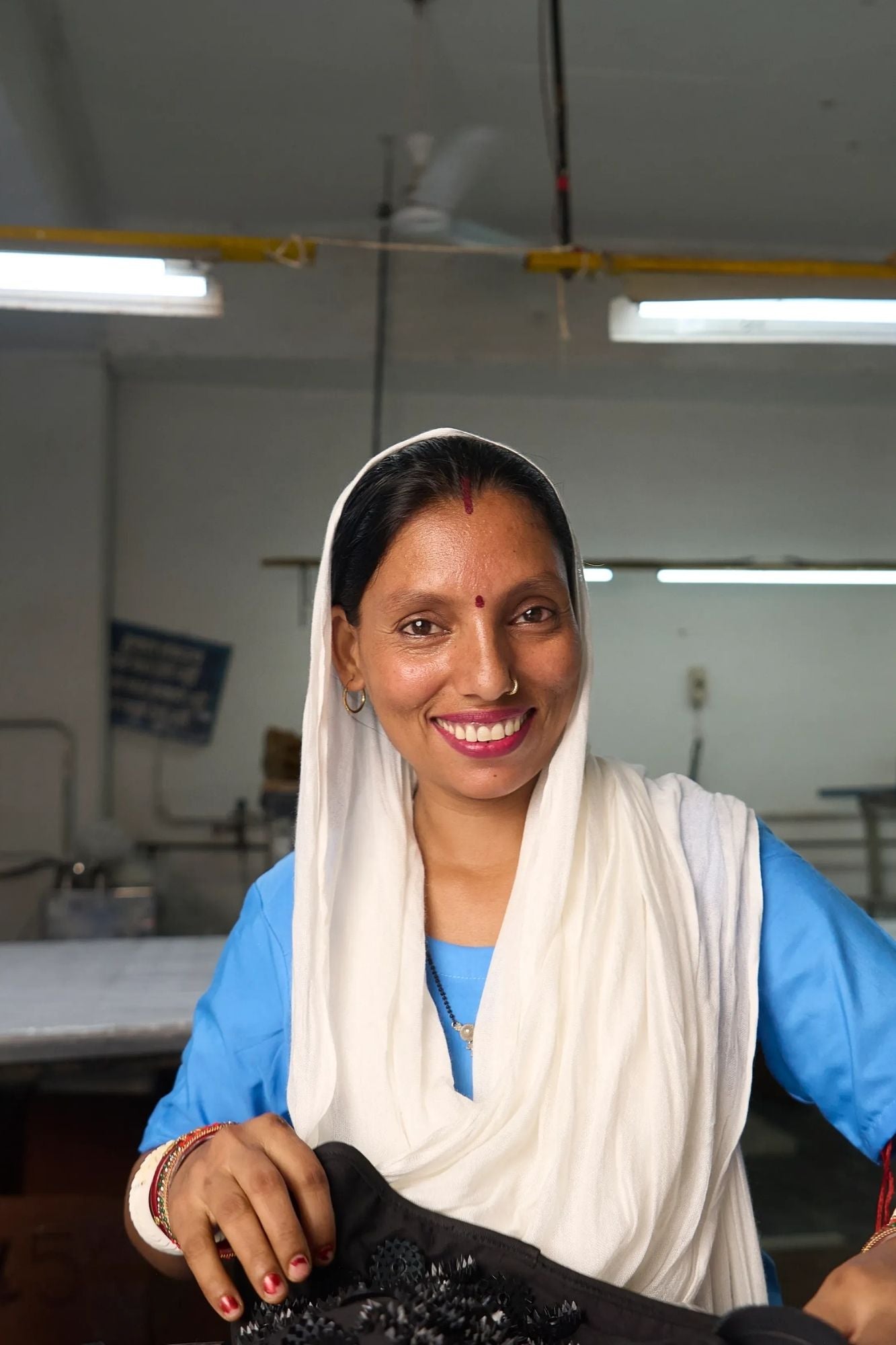 A smiling woman wearing a white headscarf and blue top holds a Shakti Leg Wrap in a workshop setting.