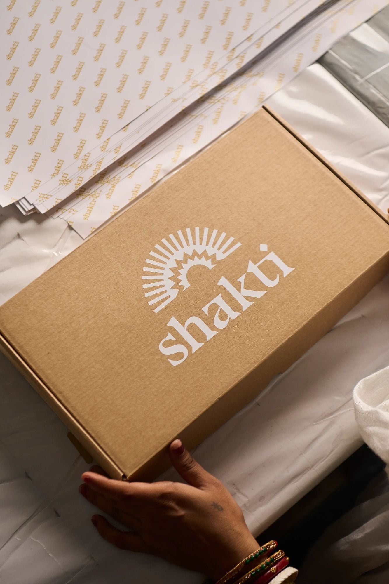 A brown cardboard box with the white Shakti logo, held by a hand over white fabric, with branded tissue paper visible in the background.